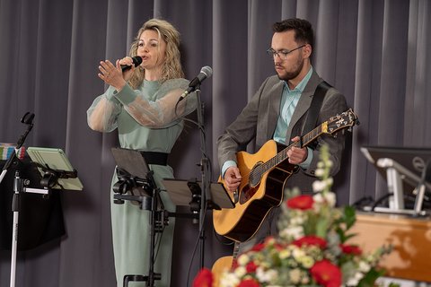 Das Musikduo "Perceived as One", die Sängerin singt ins Mikrofon, ihr Partner steht im hellem Anzug und mit dezent grünem Hemd spielt Gitarre. Links im Bild ist unscharf das Blumengesteck der Bühne zu sehen. (öffnet Vergrößerung des Bildes)