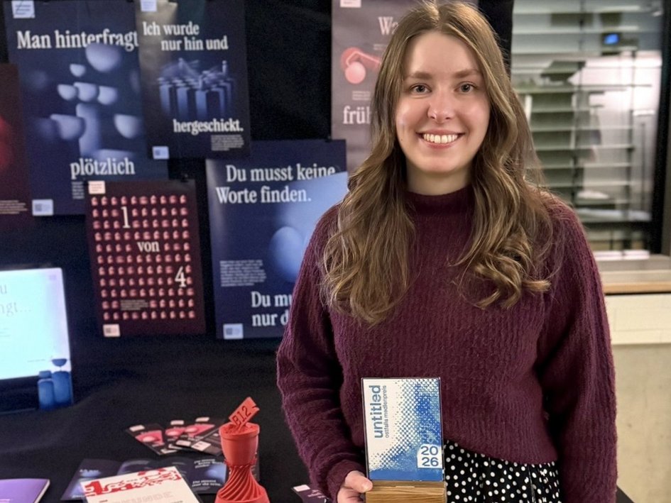 Laura Knop steht vor den Plakaten zum Thema Fehlgeburten, die sie im Rahmen ihrer Abschlussarbeit gestaltet hat. In der Hand hält sie den Ostfalia-Medienpreis
