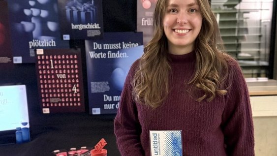 Laura Knop steht vor den Plakaten zum Thema Fehlgeburten, die sie im Rahmen ihrer Abschlussarbeit gestaltet hat. In der Hand hält sie den Ostfalia-Medienpreis