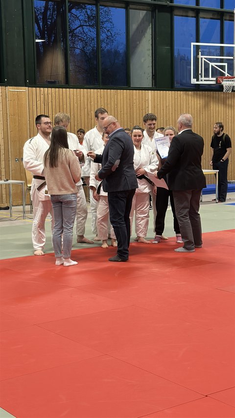 Eine Gruppe von Judoka der Ostfalia und TU Braunschweig steht barfuß auf der Matte und erhält von Offiziellen eine Urkunde während der Siegerehrung der Deutschen Hochschulmeisterschaft im Judo. (öffnet Vergrößerung des Bildes)
