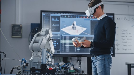 Ein Student hat eine VR-Brille auf. Im Hintergrund ist ein Bildschirm auf dem man sieht, was er sieht. Er bedient mit Joysticks einen Roboterarm