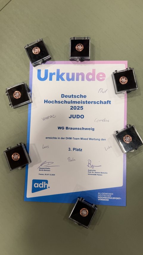 Die Urkunde für den 3. Platz der WG Braunschweig bei der DHM Judo ist auf dem Boden ausgelegt, umgeben von sieben Bronzemedaillen in kleinen Boxen. (öffnet Vergrößerung des Bildes)