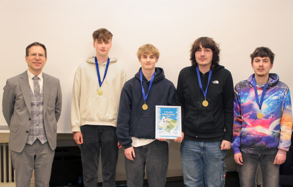 Gruppenbild von links nach rechts: Prof. Dr. Henning Zindler und vier Schüler der Sekundarstufe 2 der Wilhelm-Bracke-Gesamtschule stehen nebeneinander. Die Schüler tragen jeweils eine Medaille und halten in der Mitte das Preisgeld in einem Bilderrahmen. (öffnet Vergrößerung des Bildes)
