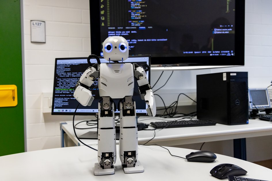 Ein Roboter steht auf einem Tisch, im Hintergrund sind Bildschirme mit Code zu sehen.