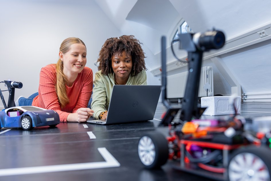 Zwei Studierende vor einem Laptop umgeben von Modellautos