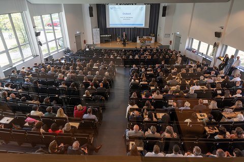 Blick von hinten auf die fast vollbesetzte Aula aus der Vogelperspektive. (öffnet Vergrößerung des Bildes)