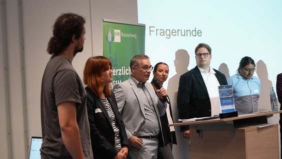 Eine Personengruppe steht vor einem Redepult. Hinter ihnen eine Leinwand mit der Aufschrift "Fragerunde"