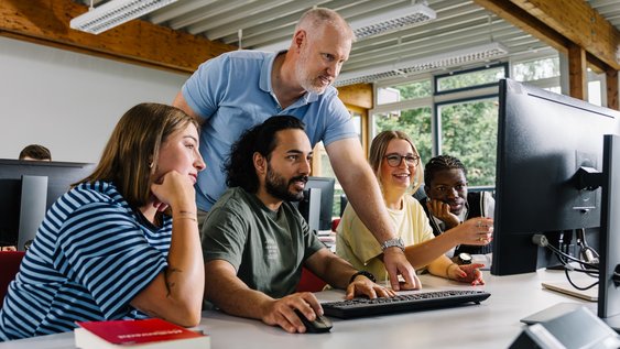 Vier Studierende sitzen vor einem PC. Ein Professor steht hinter ihnen und schaut mit ihnen gemeinsam auf den Bildschirm.