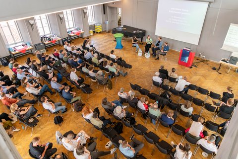 Ein Top-View-Foto des Tagungssaals. In den Reihen sitzen die Tagungsgäste, vorne sitzen die Expert*innen der Diskussionsrunde.