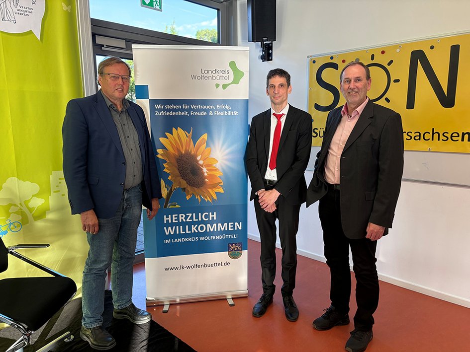 Drei Männer stehen vor einem Roll-Up des Landkreises Wolfenbüttel, das eine Sonnenblume zeigt, und lächeln in die Kamera.