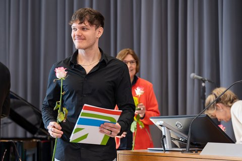 Ein Absolvent geht mit seiner Urkunde und einer Rose in der Hand lächelnd von der Bühne. (öffnet Vergrößerung des Bildes)