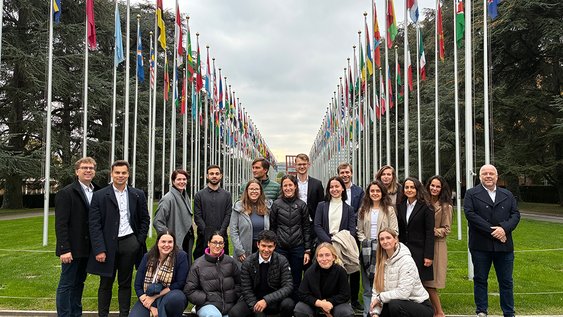 A group of about 20 people stand in front of the flags of the UN states. The people pictured are smiling into the camera.