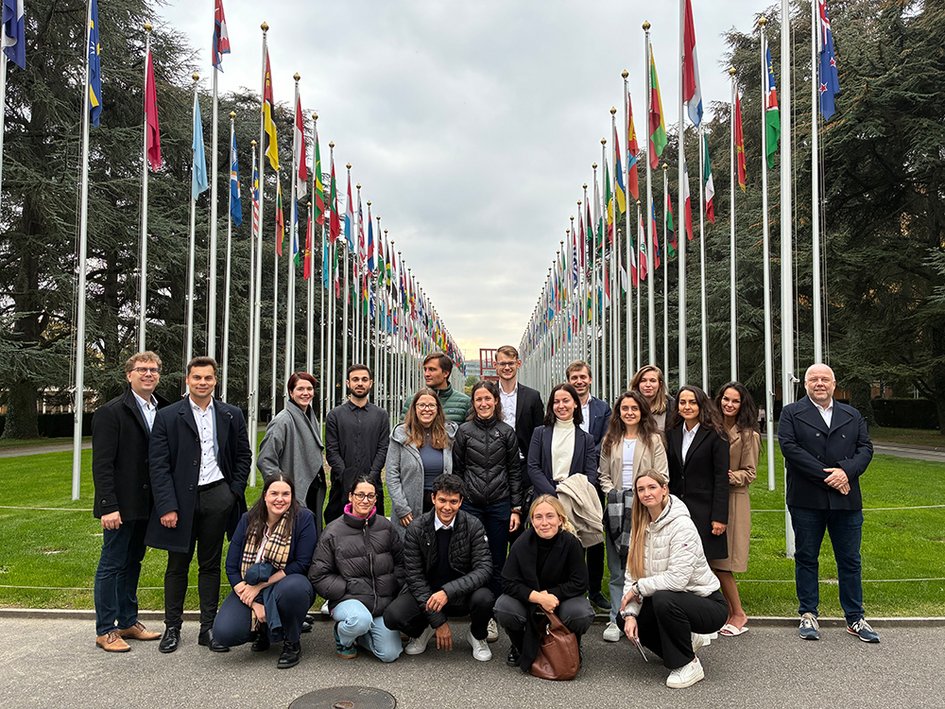 A group of about 20 people stand in front of the flags of the UN states. The people pictured are smiling into the camera.