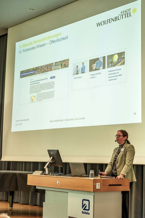 Aufnahme einer Speakerin auf der Bühne in der Aula der Ostfalia Hochschule. Im Hintergrund ist eine Präsentation an der Leinwand zum Thema Stadt Wolfenbüttel - Klimaschutz und Umwelt. (öffnet Vergrößerung des Bildes)