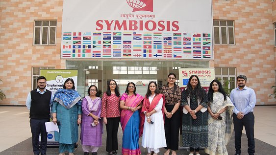 Eine Gruppe von zehn Personen vor einem Gebäude mit der Aufschrift SYMBIOSIS und etlichen Flaggen verschiedener Staaten. Die Personen lächeln in die Kamera.