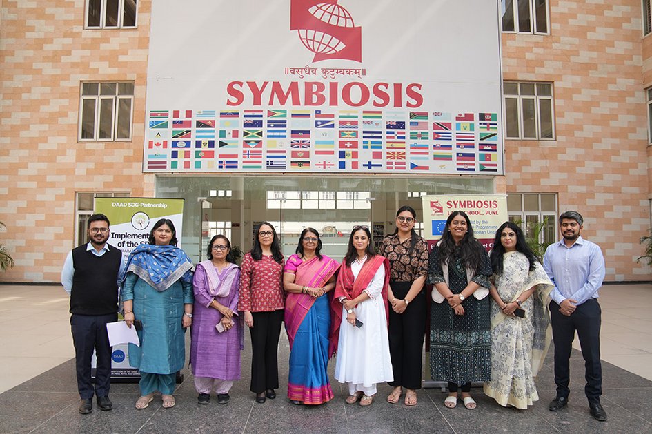 Eine Gruppe von zehn Personen vor einem Gebäude mit der Aufschrift SYMBIOSIS und etlichen Flaggen verschiedener Staaten. Die Personen lächeln in die Kamera.