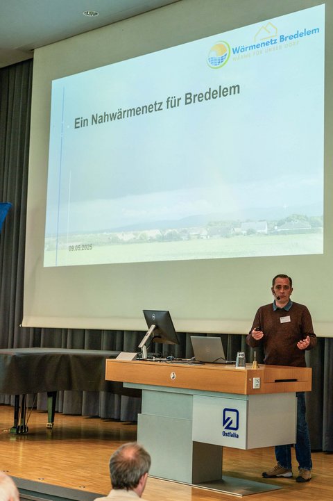 Aufnahme von der Bühne in der Aula der Ostfalia Hochschule. Auf der Bühne steht ein Speaker und erzählt über das Thema Ein Nahwärmenetz für Bredelem. Im Hintergrund ist die Präsentation dazu. (öffnet Vergrößerung des Bildes)