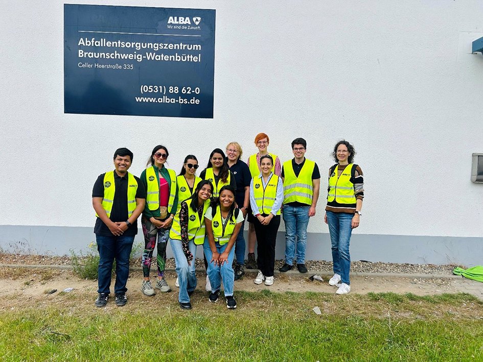 Eine Gruppe von 11 Personen steht zum Gruppenfoto vor einer Halle mit dem Schild "Abfallentsorgungszentrum Braunschweig-Watenbüttel". Die Abgebildeten tragen gelbe Warnwesten und lächeln in die Kamera