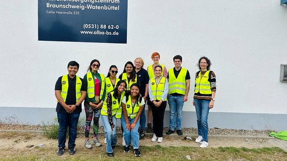Eine Gruppe von 11 Personen steht zum Gruppenfoto vor einer Halle mit dem Schild "Abfallentsorgungszentrum Braunschweig-Watenbüttel". Die Abgebildeten tragen gelbe Warnwesten und lächeln in die Kamera