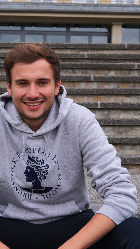 Ein junger Mann trägt den Hoodie der BELS in grau melange. Der Hoodie trägt das Logo der BELS, die Aequitas, über dem Bauch. Darum findet sich der kreisförmig angeordnete Schriftzug "Brunswick European Law School". Das Logo und der Schriftzug sind blau. (öffnet Vergrößerung des Bildes)