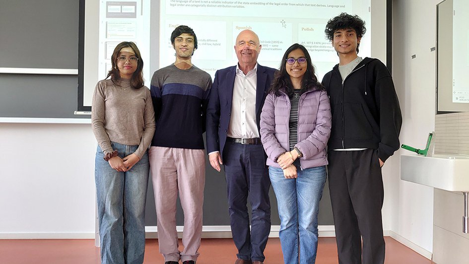A group photo showing five people standing in front of a screen on which a projector image is visible. The people in the photo are smiling at the camera.