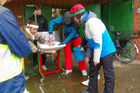 Studierende draußen vor Gebäude M befreien den Waschbeckenabfluss und tragen dabei Ski-Kostüme (öffnet Vergrößerung des Bildes)