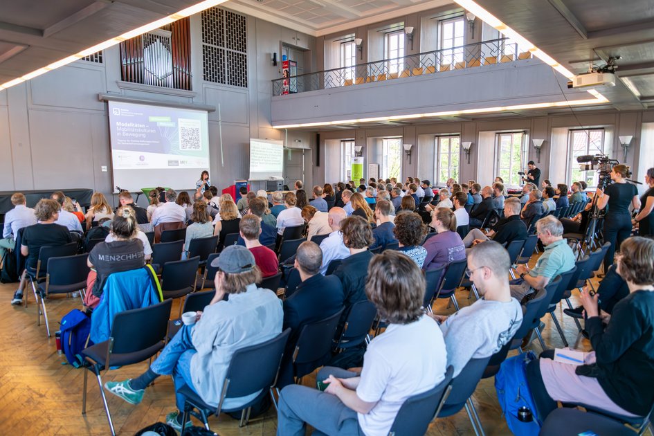 Foto vom Tagungssaal. Viele Menschen sitzen in Reihen im Publikum. Man sieht das Publikum in Rückenansicht und vorne im Saal die Moderation, die die Tagung eröffnet.