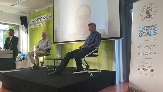 Prof. Litschen steht links am Rednerpult, Hans-Heinrich Schmidt-Kanefendt, Benjamin Volz (digital zugeschaltet), und Prof. Dr. Jürgen Kuck diskutieren miteinander, Benjamin Volz ist auf einer Leinwand zu hinter der Bühne zu sehen.