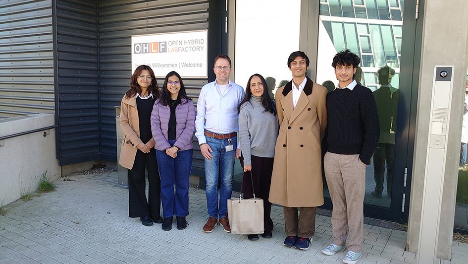 A group photo of six people in front of the entrance to the Open Hybrid LabFactory. The people in the photo are smiling at the camera.