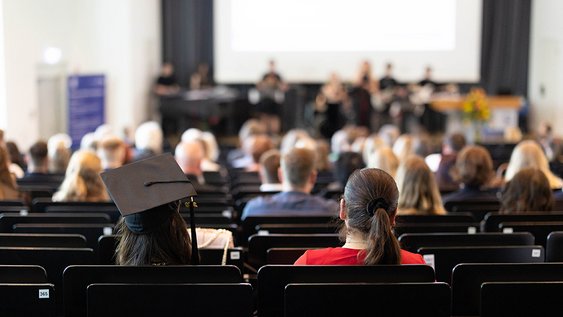 Eine Person mit Graduation-Hut sitzt mit anderen im Auditorium der Aula, Musiker auf der Bühne sind verschwommen zu sehen.