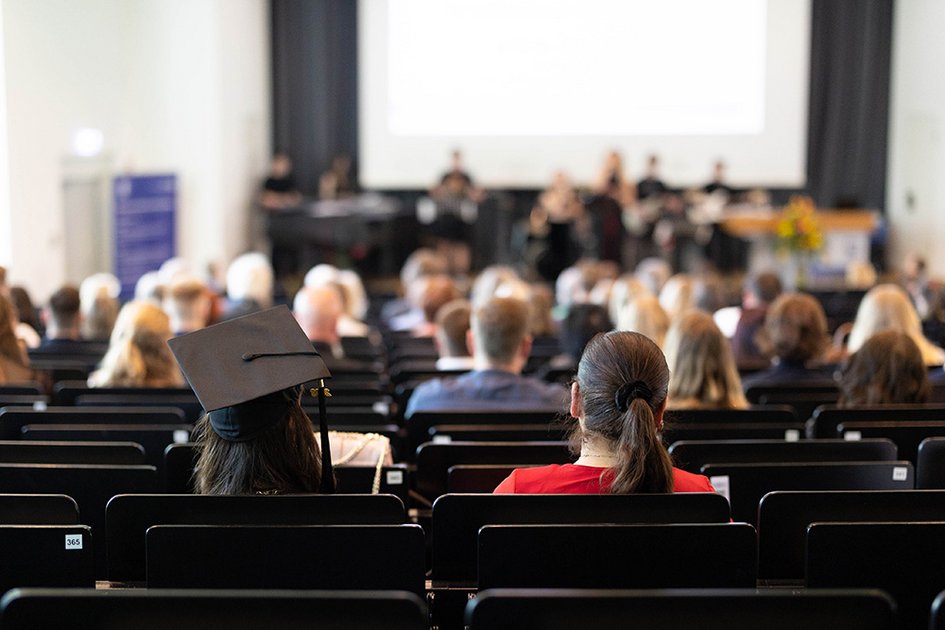 Eine Person mit Graduation-Hut sitzt mit anderen im Auditorium der Aula, Musiker auf der Bühne sind verschwommen zu sehen.