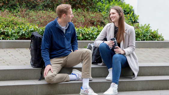 Zwei Studierende sitzen auf der Treppe im Außengelände und unterhalten sich.