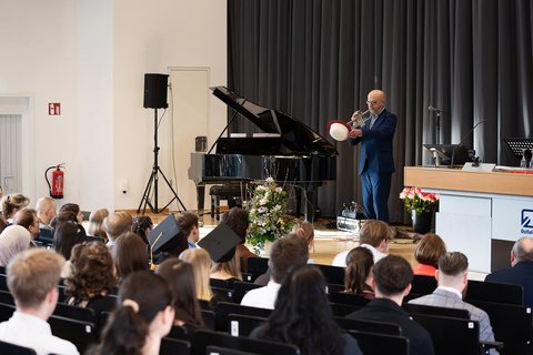 Géza Gál, Leiter der TU Big Band Braunschweig, steht auf der Bühne und spielt mit einer Hand Trompete, während er mit der anderen einen Hut hält. (öffnet Vergrößerung des Bildes)