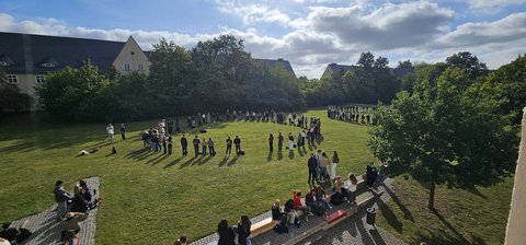 Auf einer Wiese stehen viele Studierende im Kreis. (öffnet Vergrößerung des Bildes)