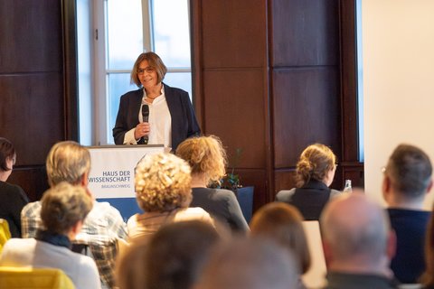 Prof. Dr. Sandra Verena Müller steht vorne am Pult und hält ein Mikrofon in der Hand und spricht zum Publikum. (öffnet Vergrößerung des Bildes)