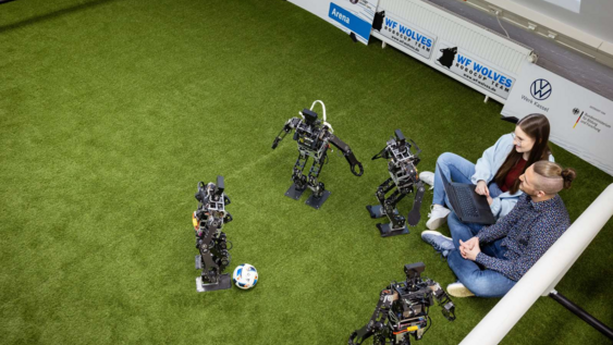 2 Studierende sitzen auf dem Biden und arbeiten mit 4 Fussballrobotern