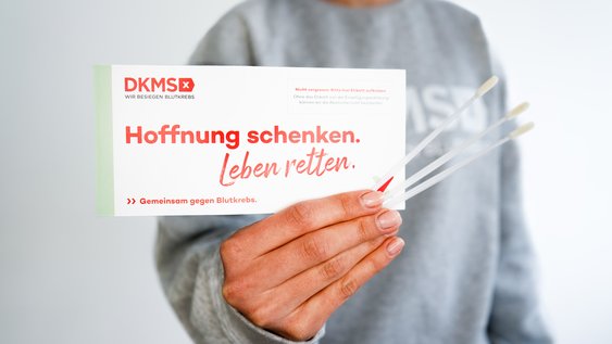 Jemand hält einen Zettel mit den Worten "Hoffnung schenken. Leben retten" in der Hand sowie Wattestäbchen für die DKMS-Registrierung