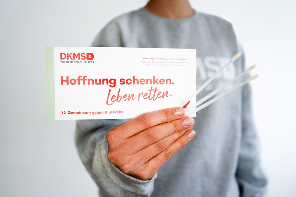 Jemand hält einen Zettel mit den Worten "Hoffnung schenken. Leben retten" in der Hand sowie Wattestäbchen für die DKMS-Registrierung
