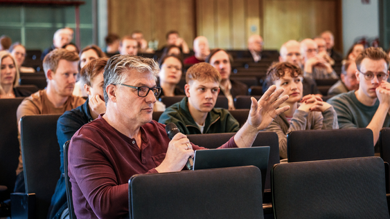 Zuschauer der Fachtagung 2025 sitzen in der Aula. Im Vordergrund hält ein Zuschauer das Mikrofon und spricht hinein.