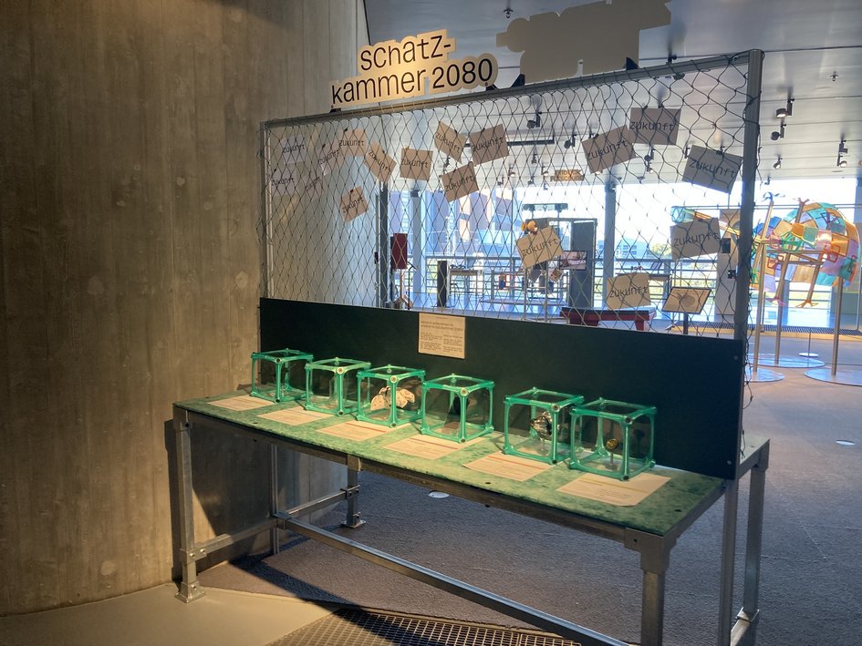 Ausstellungsstation „Schatzkammer 2080“ mit beschrifteten Zukunftskarten an einem Netz und mehreren transparenten Schaukästen mit Gegenständen auf einem Tisch.