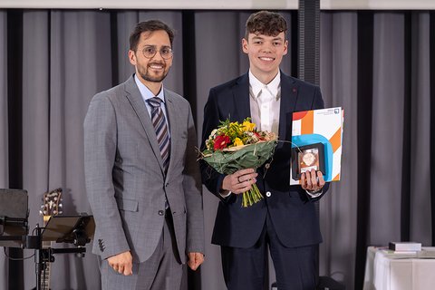 Rechtsanwalt Adrian Linden im grauen Anzug neben dem Preisträger Marc Michaelis. Der Ausgezeichnete im blauen Anzug hält den Eckert & Kollegen Award 2026 und einen Blumenstrauß. Beide lächeln in die Kamera. (öffnet Vergrößerung des Bildes)