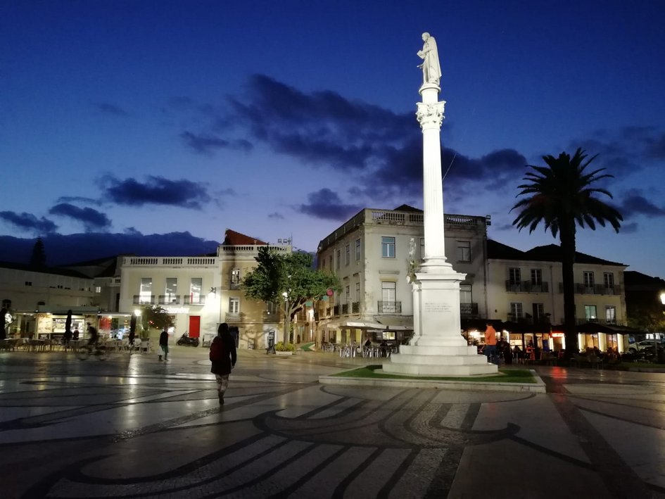 Eine Fotografie des Bocage-Platz in der portugiesischen Stadt Setúbal bei Nacht. Im Fokus steht eine beleuchtete Statue auf einer weißen Säule