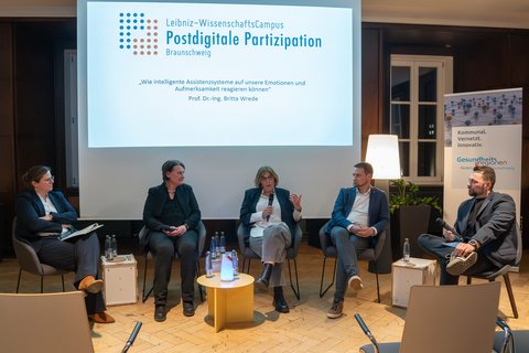 Ein Blick auf die Podiumsdiskussion, welche vorne, vor der Beamer Leinwand statt fand, auf welcher das Logo des WissenschaftsCampus und der Titel des Vortrags angezeigt werden. Die Teilnehmer:innen sitzen auf Stühlen im Halbkreis, um einen kleinen, runden Tisch. (öffnet Vergrößerung des Bildes)