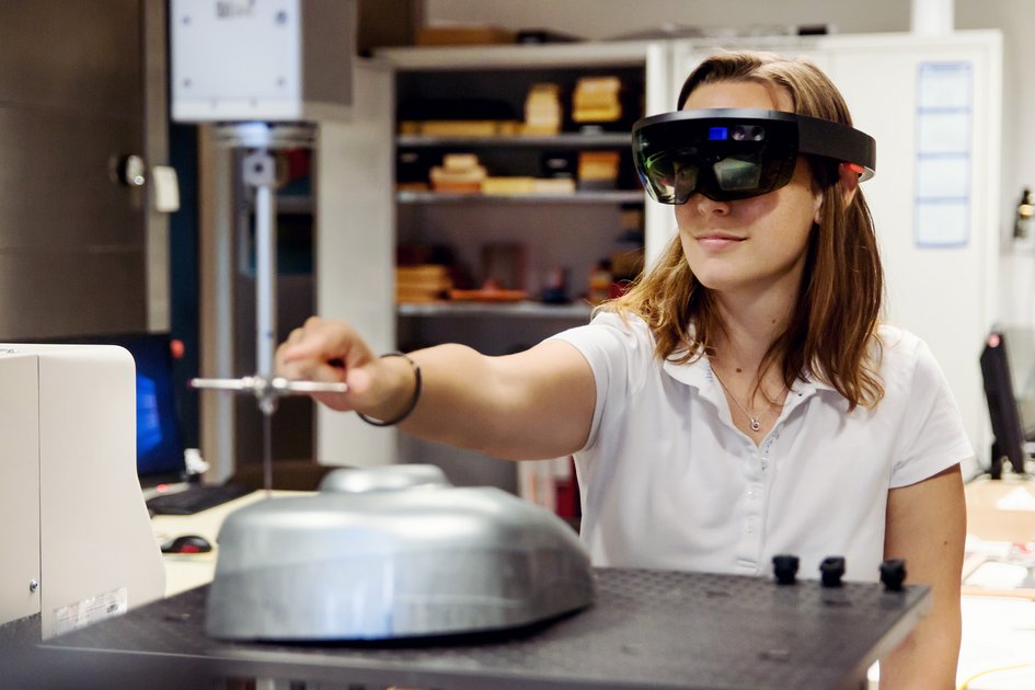 Eine Frau steht in einer Industriehalle und trägt eine AR-Brille. Sie streckt ihre Hand aus und interagiert mit einem Objekt, das sie durch ihre Brille sieht.