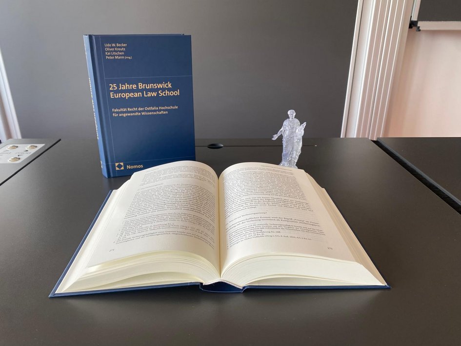 Das Bild zeigt die Festschrift ein Malstehend, ein Mal aufgeschlagen auf dem einem Tisch in einem Vorlesungsraum. Rechts neben den Festschriften steht eine kleine Figurine der Aequitas.