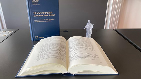 Das Bild zeigt die Festschrift ein Malstehend, ein Mal aufgeschlagen auf dem einem Tisch in einem Vorlesungsraum. Rechts neben den Festschriften steht eine kleine Figurine der Aequitas.