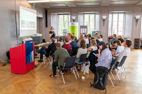 Foto einer Präsentation: Vorne referiert eine Wissenschaftlerin während in einem Halbkreis in mehreren Reihen Zuschauende sitzen.