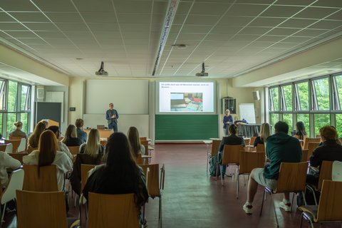 Vorlesungsraum voller Schüler*innen, die Richtung Beamerleinwand sitzen, die ein Bild mit Laptop und Büchern zeigt, mit der Bildüberschirft "Aber ich wollte doch was mit Menschen studieren...". Daneben Karl-Heinz-Gröpler und Christine Jahn, welche gerade Fragen beantworten. (öffnet Vergrößerung des Bildes)
