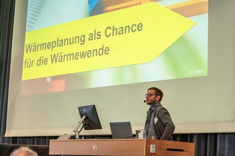 Nahaufnahme von der Bühne. Ein Speaker steht auf der Bühne und erzählt über das Thema Wärmeplanung als Chance für die Wärmewende. (öffnet Vergrößerung des Bildes)