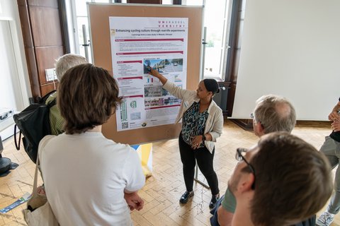 Foto einer Postersession: Eine Gruppe steht vor einer Stellwand auf der ein Plakat zu sehen ist. 